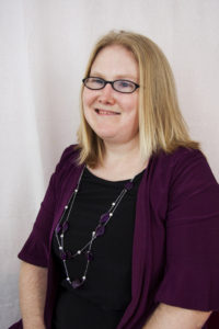 A headshot of a white woman smiling with shoulder length blonde hair, wearing glasses, a black top and purple cardigan, and long necklace. 