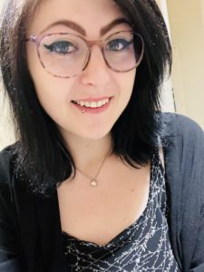 A head shot of a white woman smiling with shoulder length black hair, glasses, a black and white top and black cardigan, necklace, and lip ring.