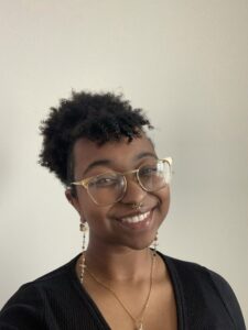 A headshot of a black woman, smiling and wearing glasses, with short black hair pulled back in a bun, dangly earrings, nose ring, a necklace, and a black top.